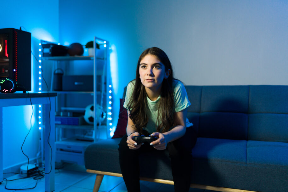 young woman sitting at the couch in her bedroom with led lights while holding a remote controller and playing a video game from a gaming pc.